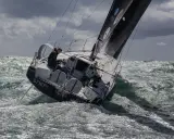 Xavier Macaire navigue sur un voilier dans une mer agitée, le bateau est incliné, les vagues déferlent.