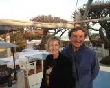 Patrice de Colmont et une femme souriants sur une terrasse de plage, avec des arbres dénudés et des sièges blancs en arrière-plan.