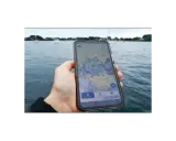 Main d'une personne tenant un smartphone affichant une carte marine avec des points d'intérêt. Le nav et co. sont visibles sur l'eau.