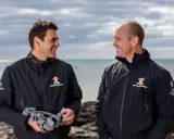 Damien Guillou et son collègue souriants sur la plage, l'un tenant un sextant, prêts pour une nouvelle aventure maritime.