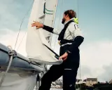 Un homme ajuste un type de voile sur un voilier, avec des maisons en arrière-plan.