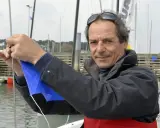 Marc Guillemot, skipper expérimenté, ajuste une voile bleue sur un ponton, prêt pour une nouvelle aventure en mer.