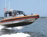 Un bateau du garde côte américain file sur l'eau, créant une gerbe d'écume. Deux membres d'équipage sont visibles à l'intérieur.