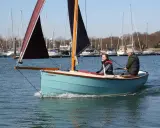 Deux hommes naviguent sur un petit bateau à voile bleu clair, avec d'autres voiliers en arrière-plan.
