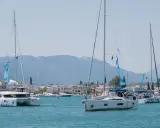 Trois voiliers naviguent dans une baie ensoleillée. Au premier plan, un catamaran monocoque blanc avec des voiles bleues et blanches. Derrière, un autre voilier blanc avec des personnes à bord. Au loin, une ville côtière et des montagnes.