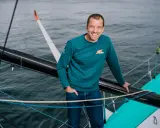 Sam Goodchild, souriant, sur un voilier, portant un sweat "For the Planet".