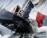 Le voilier Pen Duick VI fend les vagues, pavillon français au vent. Une silhouette se tient à la barre.