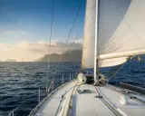 Le voilier navigue sur une mer bleue, le grand voile blanc gonflé par le vent. Au loin, des îles brumeuses se dessinent sous un ciel clair.