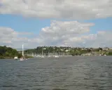Vue d'un port de plaisance, avec des voiliers amarrés et des bateaux à moteur naviguant. On aperçoit une ville en arrière-plan, perchée sur une colline verdoyante, et un pont au loin. L'ambiance est paisible, propice à une balade pour remonter la vilai...