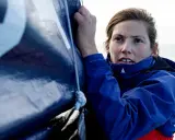 Tiphaine Ragueneau, concentrée, ajuste une voile bleue sur un bateau. La mer est visible en arrière-plan.