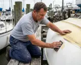 Un homme ponce le flanc d'un bateau pour refaire le gelcoat. Il est à genoux sur un ponton, entouré d'autres voiliers.