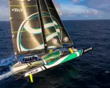 Un trimaran aux voiles noires et blanches, arborant le logo "sails of change", fend les vagues bleues sous un ciel nuageux.