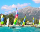 Plusieurs Hobie Cat 16 naviguent sur une mer turquoise, voiles multicolores, montagne en arrière-plan.