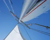 Vue plongeante sur le mat de bateau, voiles blanches déployées sous un ciel bleu éclatant, cordages tendus.