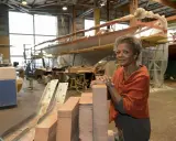 Jacqueline Tabarly, souriante, dans un atelier de construction navale, devant des blocs de bois et un voilier en cours de fabrication.