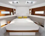 L'intérieur d'un voilier 8m, avec un grand lit blanc, des coussins jaunes, des hublots donnant sur la mer et des étagères en bois.
