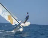 Un homme en trapeze sur un catamaran navigue sur une mer bleue sous un ciel clair. La voile arbore un dessin original.