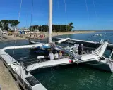 Le trimaran Flo, un bateau à trois coques, est amarré à quai. Des personnes sont à bord et sur la plage en arrière-plan.