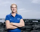 Rosalin Kuiper, souriante, pose les bras croisés devant un paysage côtier rocheux.