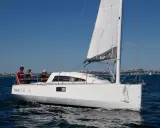 Un voilier Ikone 7.50 navigue par beau temps, avec trois personnes à bord. La voile est hissée, le ciel est bleu.