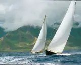 Deux voiliers de course, dont un avec le numéro 7020, naviguent sur une mer agitée, avec des montagnes verdoyantes et brumeuses en arrière-plan. Le pen duick 3 est en pleine action.
