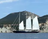 Le yacht de Bezos, un voilier majestueux, navigue en Méditerranée, ses voiles blanches déployées devant des falaises escarpées.