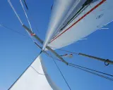 Vue plongeante sur le mât et les voiles d'un voilier, le ciel bleu intense en arrière-plan. L'image capture l'essence de la navigation, le vent gonflant les voiles.