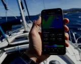 Sur un voilier, une main tient un smartphone affichant une application météo marine gratuite avec les prévisions pour Ibiza.