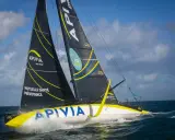 Le voilier Apivia, skippé par Alex Thomson, fend les vagues sous un ciel bleu et nuageux.