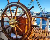 Roue de gouvernail en bois et laiton sur le pont d'un bateau. En arrière-plan, des yachts modernes et des bâtiments dans un port ensoleillé.