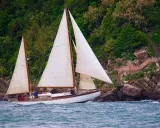 Un magnifique voilier 2 mâts navigue près d'une côte rocheuse verdoyante.