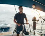 Un homme souriant barre un bateau, profitant du soleil couchant. Un petit bateau gonflable est attaché à l'arrière.