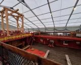 L'intérieur du **bateau Hermione** est en cours de restauration. On voit la structure en bois, les ponts rouges et jaunes, et un canon.