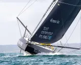 Le voilier IMOCA de Jean Le Cam fend les vagues, sa grand-voile noire ornée du slogan "Tout commence en Finistère" gonflée par le vent.
