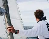 Un jeune homme regarde l'horizon depuis un voilier autonome. La voile blanche est gonflée par le vent.