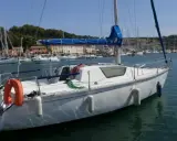 Bateau blanc à quai, voiles bleues enroulées. Au loin, un port animé avec des maisons colorées et d'autres voiliers. Un endroit idéal pour un séjour, comme le suggèrent les gib sea 76 avis.