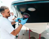 Un homme polit la coque d'un bateau. Il utilise une polisseuse pour un travail de précision, peut-être en prévision d'une révision moteur inboard.