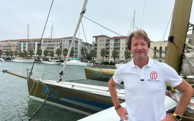 Un homme souriant sur un trimaran, peut-être le "Florence Arthaud", avec des bateaux et des immeubles en arrière-plan.