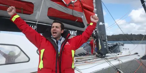 Un marinier en veste rouge célèbre sur un voilier. Une opportunité unique : ce voilier est à donner gratuitement !