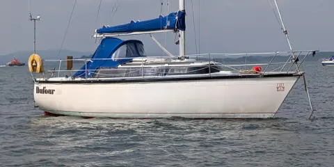 Voilier blanc Dufour 2800 à l'ancre, avec une bâche bleue sur la cabine. Un remorqueur orange est visible au loin.
