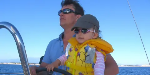 Un homme, le chef de bord bateau, tient son enfant sur ses genoux, naviguant sous un ciel bleu.