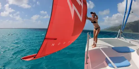 Une femme en bikini ajuste la voile rouge d'un petit catamaran habitable. L'eau turquoise et le ciel bleu créent un décor paradisiaque.