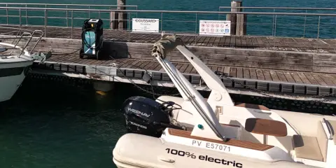 Un bateau électrique est prêt à être rechargé sur un ponton en bois. Une borne de recharge AQUA est visible à proximité.