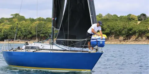 Un homme prépare le bateau, une voile noire déployée, sur fond de côte boisée. Il semble être le **half toner** de l'équipage.