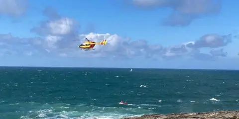 Un hélicoptère jaune et rouge survole une mer agitée, cherchant une personne disparue en mer. Un petit bateau rouge est visible au loin.
