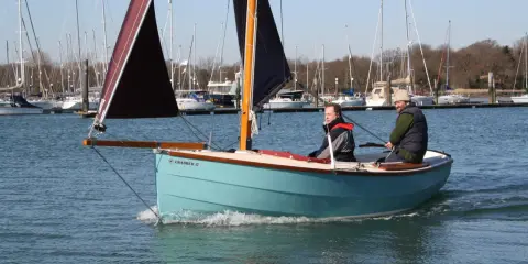 Deux hommes naviguent sur un petit bateau à voile bleu clair, avec d'autres voiliers en arrière-plan.