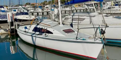 Un voilier blanc avec une voile bleue dans un port, reflété dans l'eau calme.