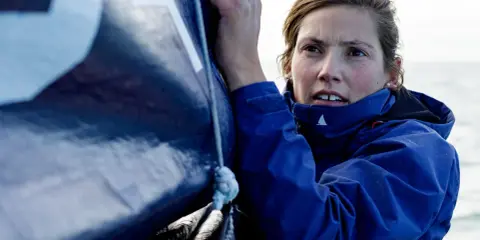 Tiphaine Ragueneau, concentrée, ajuste une voile bleue sur un bateau. La mer est visible en arrière-plan.