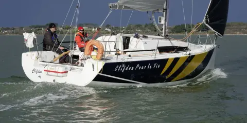 Voile rapide, le voilier "Clifden Pour la Vie" navigue sur une mer agitée. Deux personnes sont à bord, profitant du soleil.