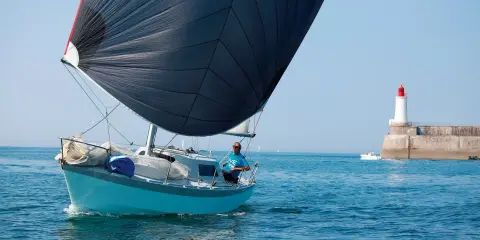 Un voilier bleu avec une grande voile noire navigue près d'un phare. Le skipper, portant un t-shirt bleu, semble observer un ovni 25 dans le ciel clair.